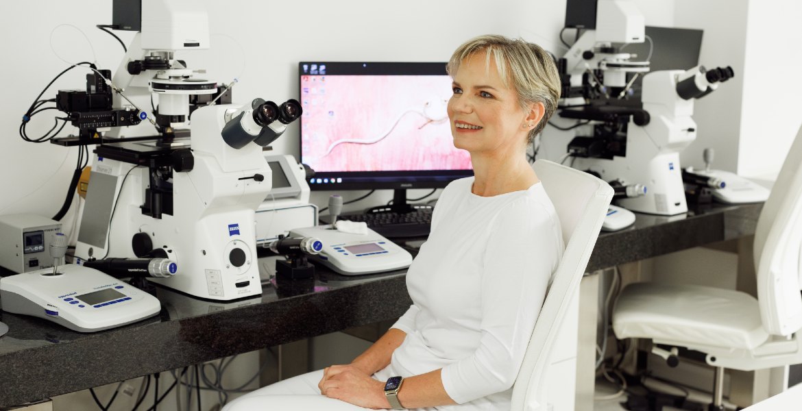 MSc Renata Hüttelova in an IVF laboratory, seated beside microscopes and fertility testing equipment, Zeiss branded devices visible on desk.
